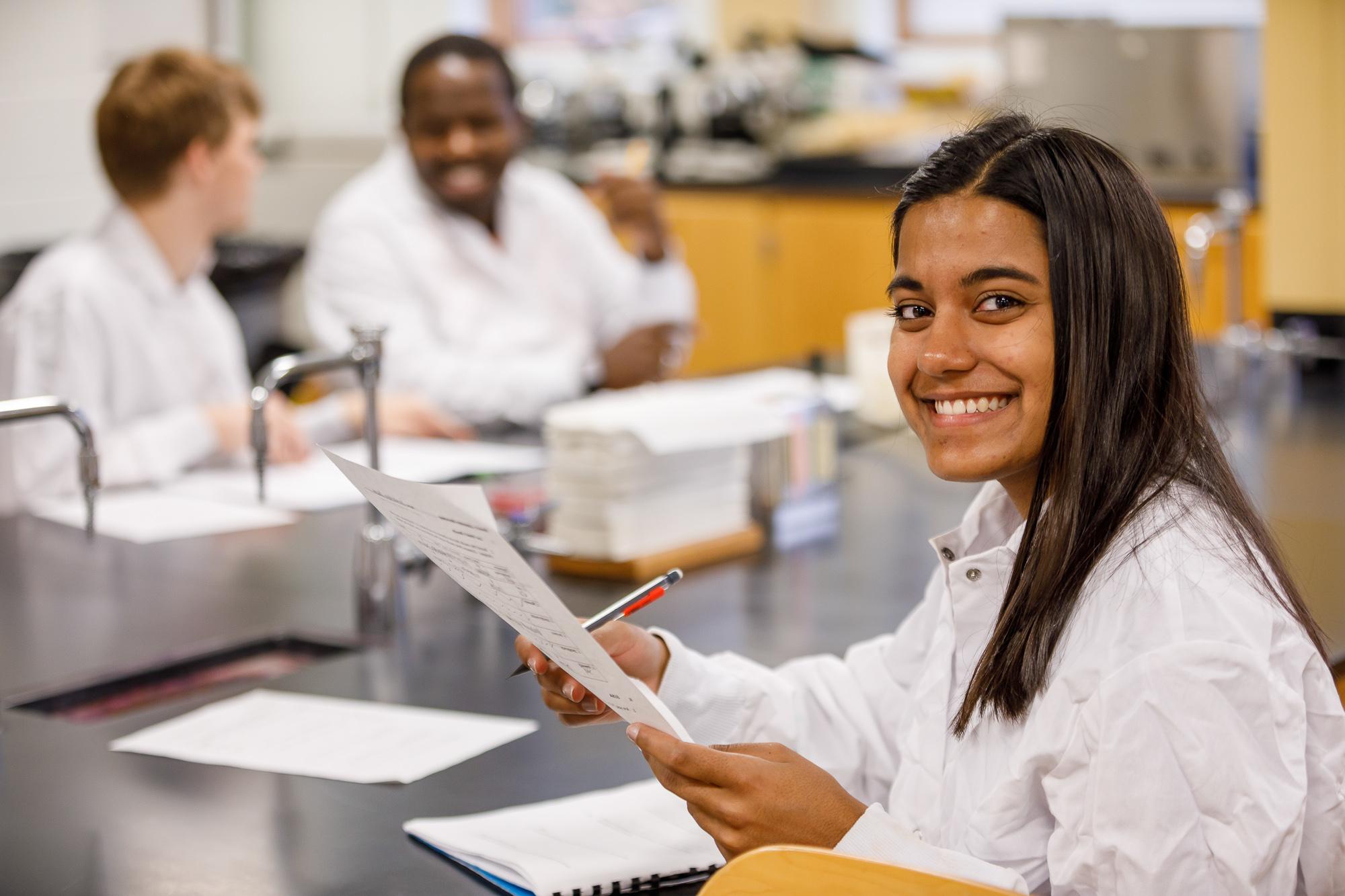 一名身穿实验服的女子面带微笑，手里拿着一篇论文，两个学生在她身后交谈